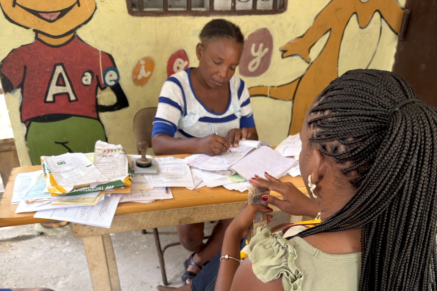 A school registration desk in Cap-Haïtien where a clerk records a payment in the school ledger while an HCTF coordinator counts out the cash.