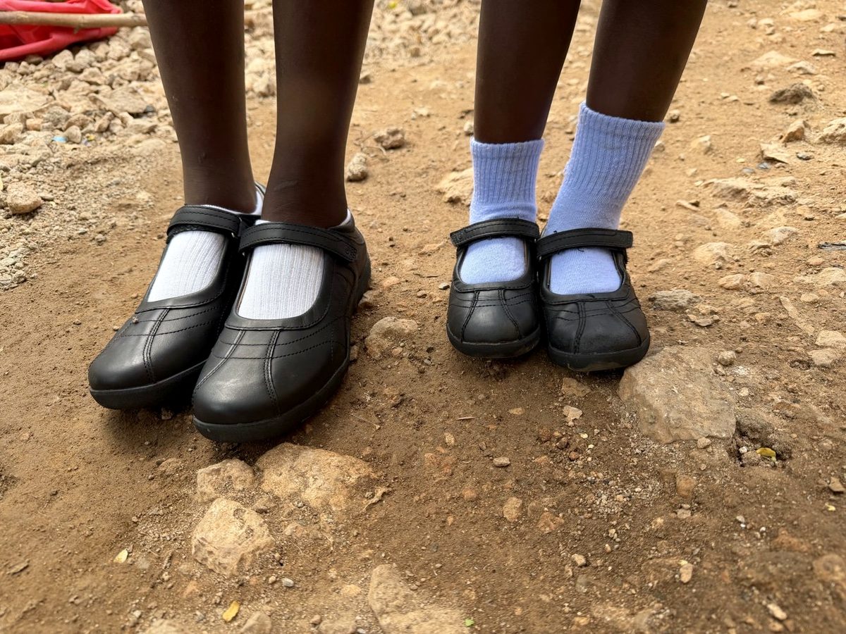 Two children stand on a dirt path in new black school shoes and socks.